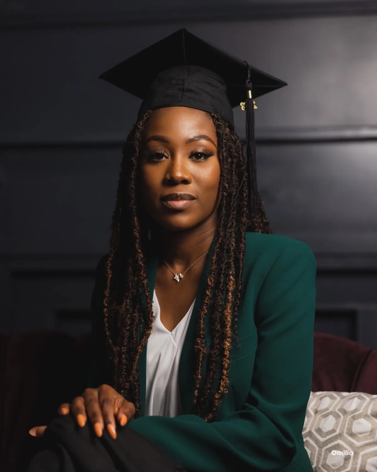 Woman wearing graduation cap and green blazer seated against dark panelled backdrop, calm expression