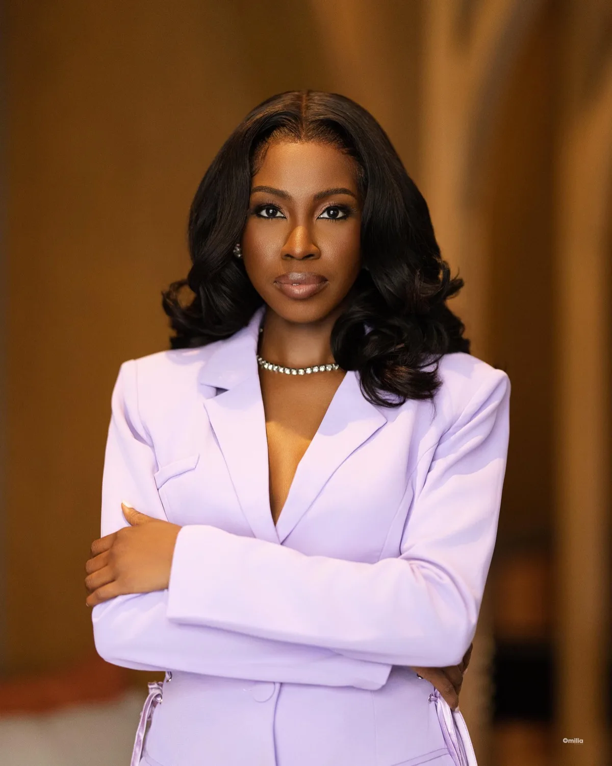 Woman in lilac blazer with arms crossed and confident expression against a warm blurred background