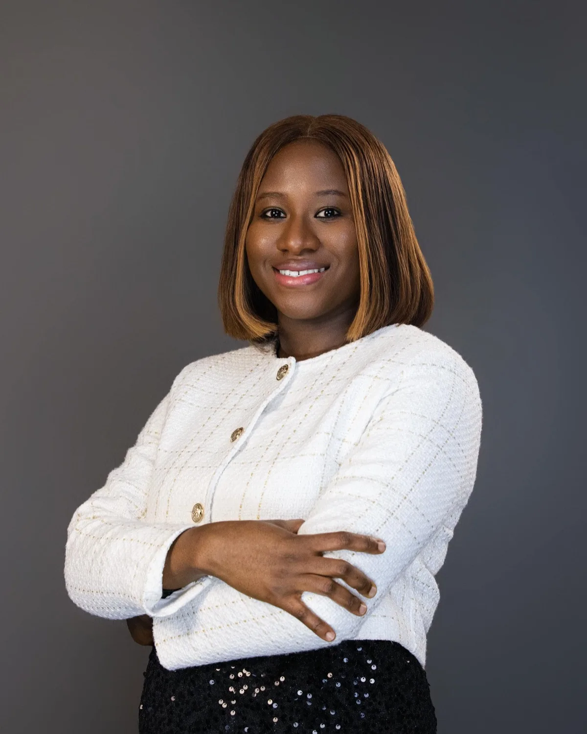 Smiling woman in white tweed jacket with gold buttons, arms crossed, against grey studio backdrop