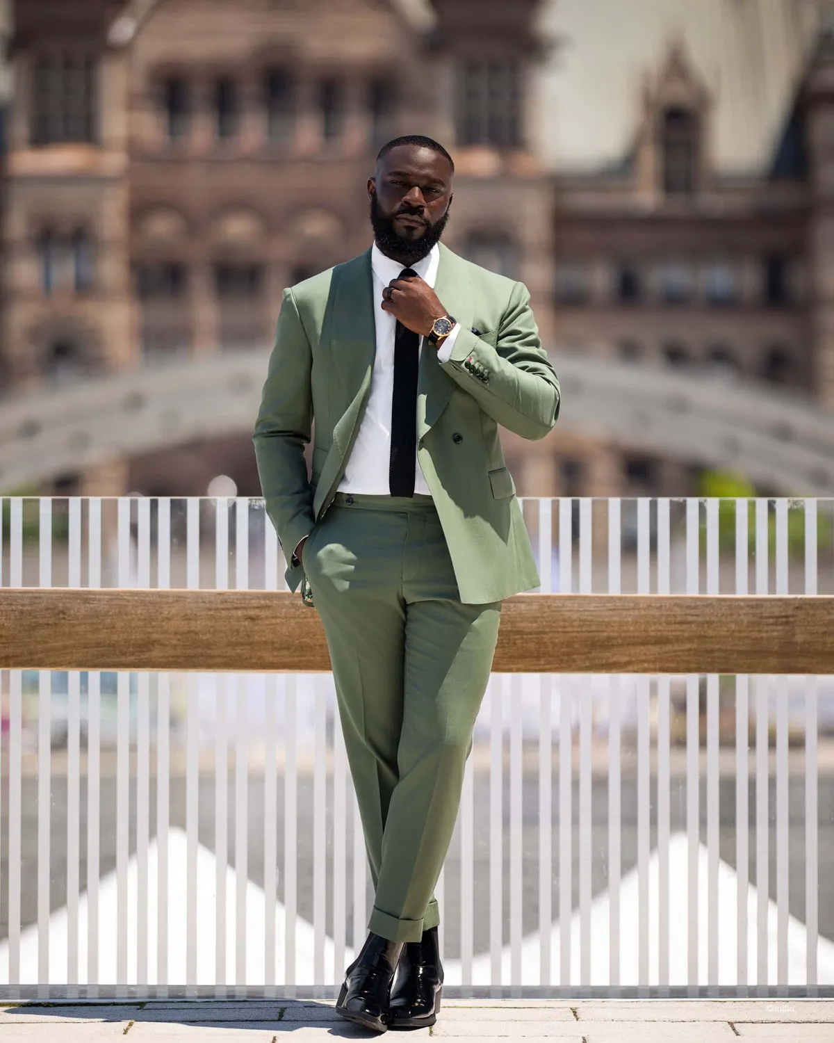 Man in sage green suit and black tie adjusting collar by railing before ornate historic building