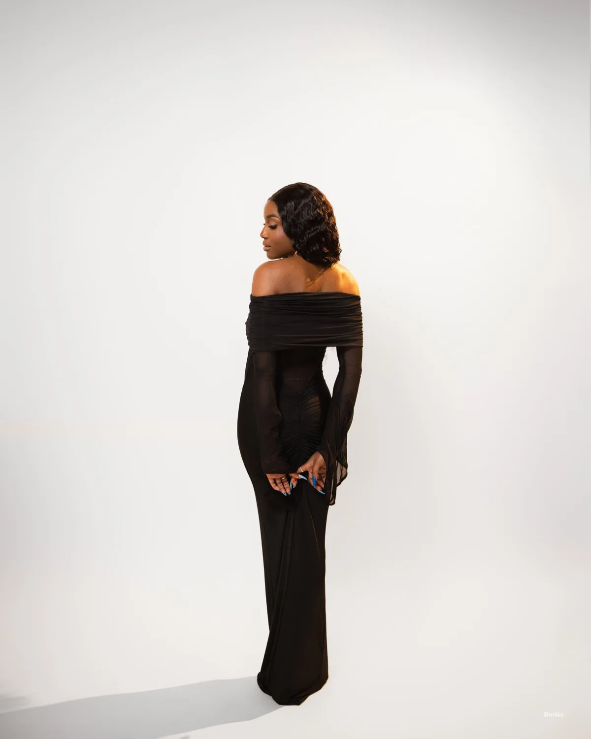 Woman in long black off-shoulder gown standing with back turned against bright white backdrop