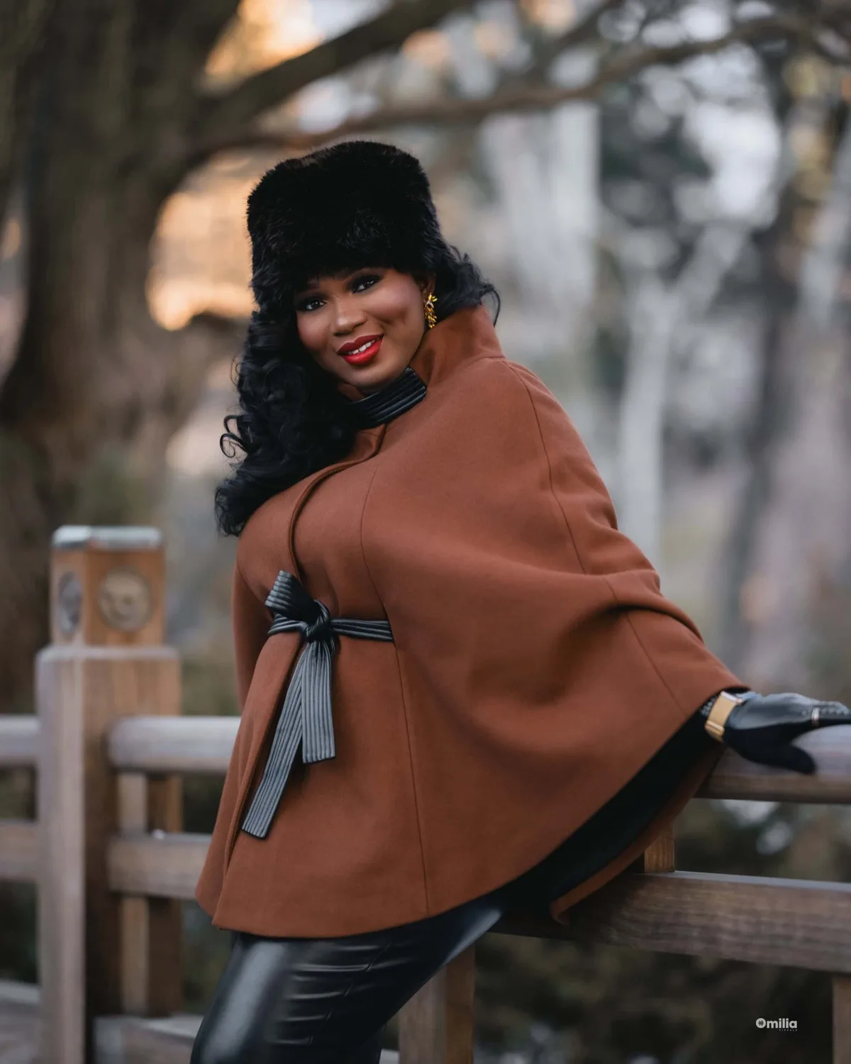 Woman in camel cape coat and black fur hat leaning on wooden fence outdoors at dusk