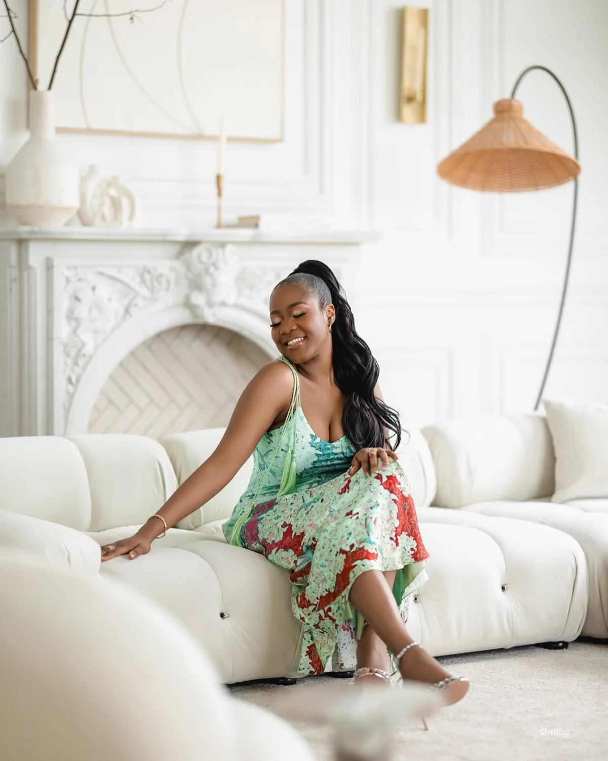 Woman in green floral dress seated on white tufted sofa smiling in bright elegant living room