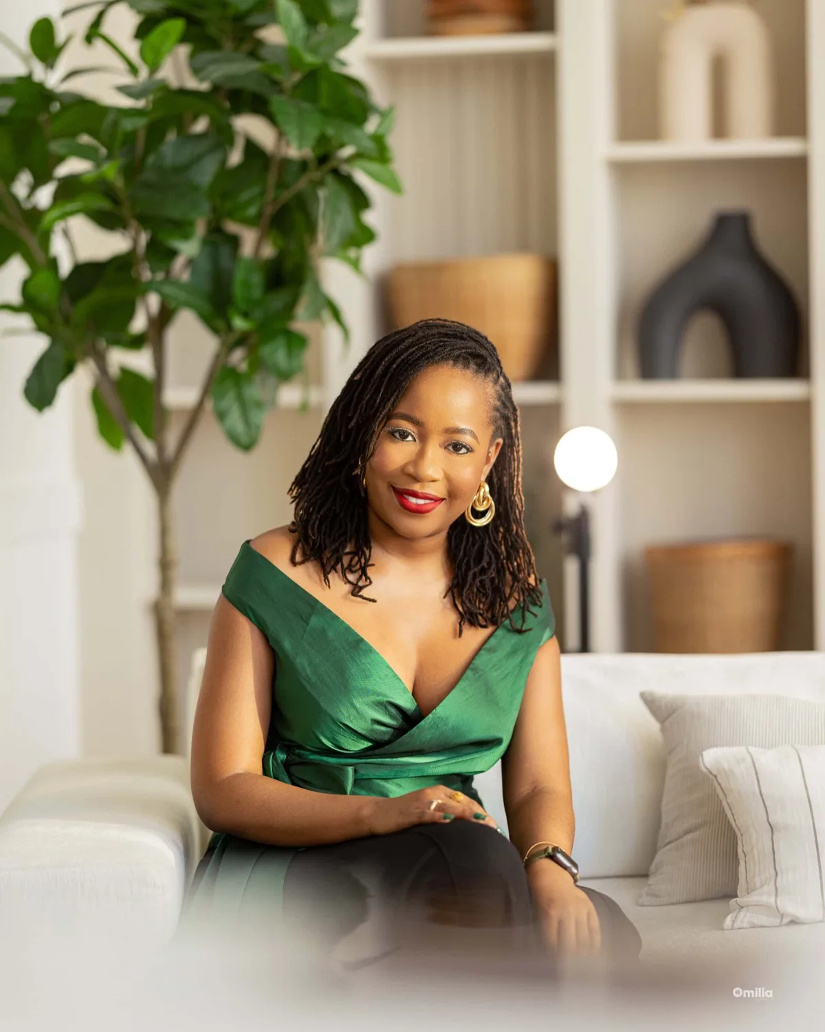 Smiling woman with shoulder-length locs in green off-shoulder top seated on white sofa in styled room