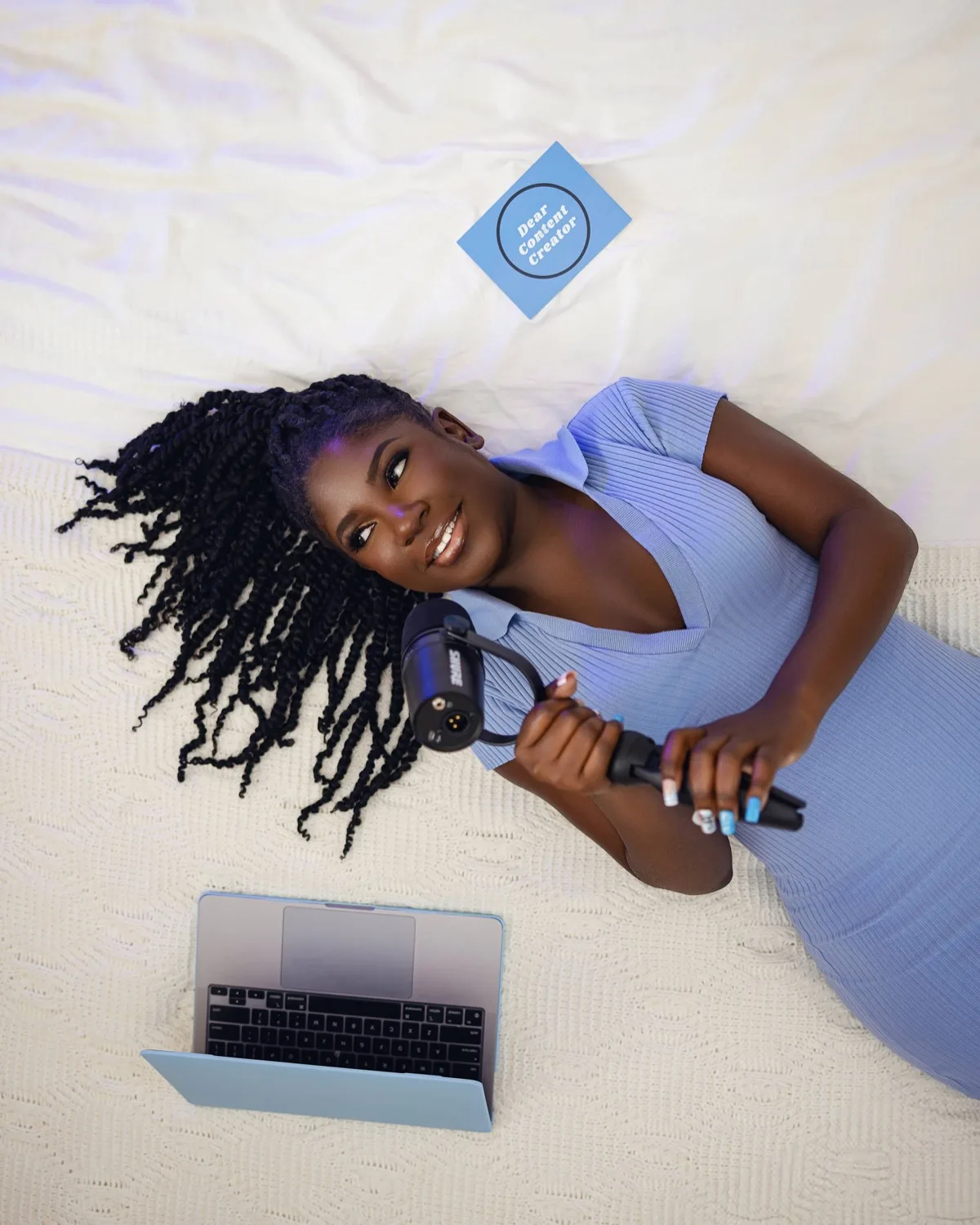 Overhead view of woman in blue dress lying down holding microphone beside open laptop