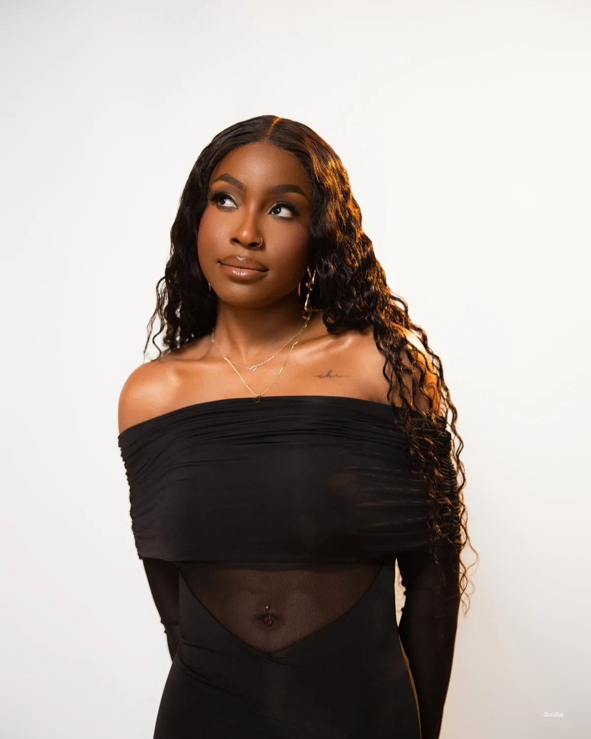 Young woman with long curly hair in off-shoulder black mesh top against soft cream backdrop