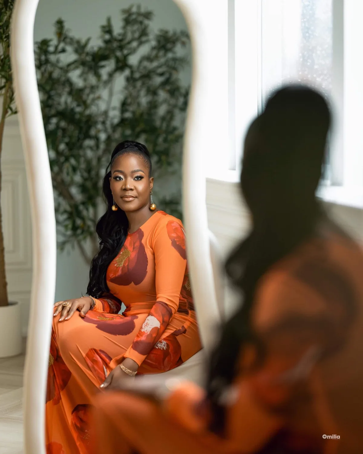 Woman in orange floral dress seated, framed by a wavy white mirror beside a leafy plant