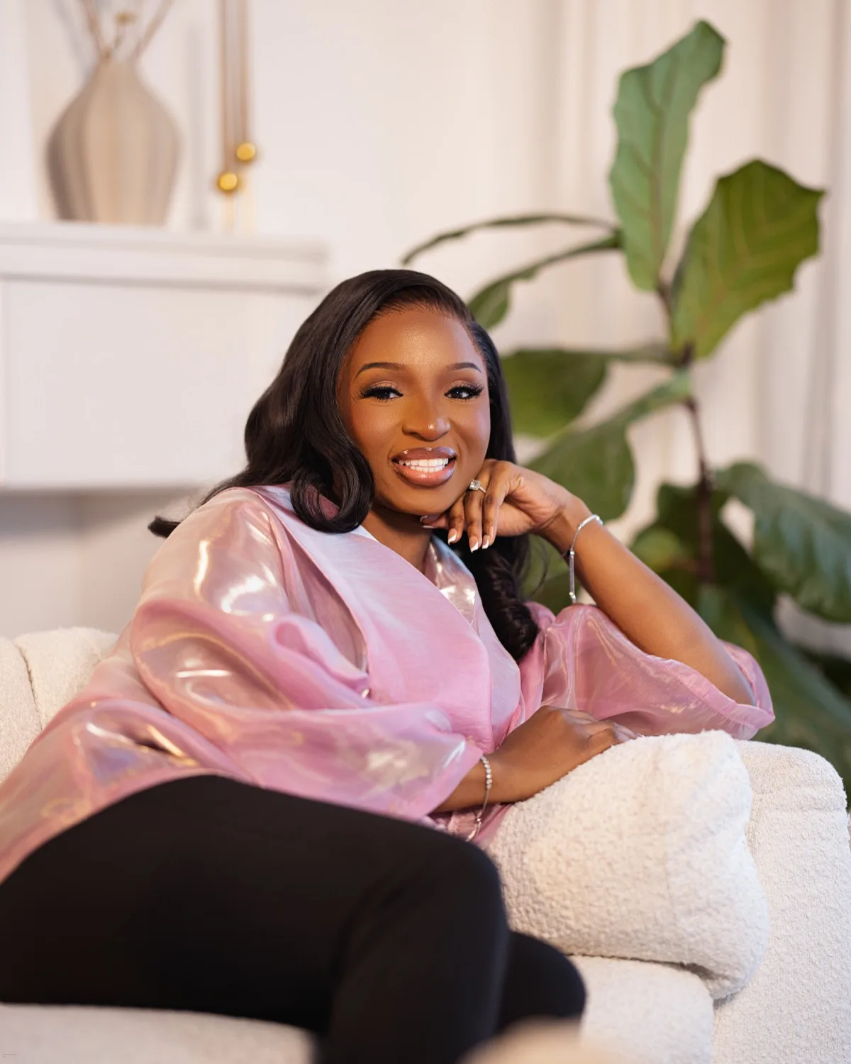 Smiling woman in iridescent pink blouse relaxing on a white sofa beside a leafy plant indoors