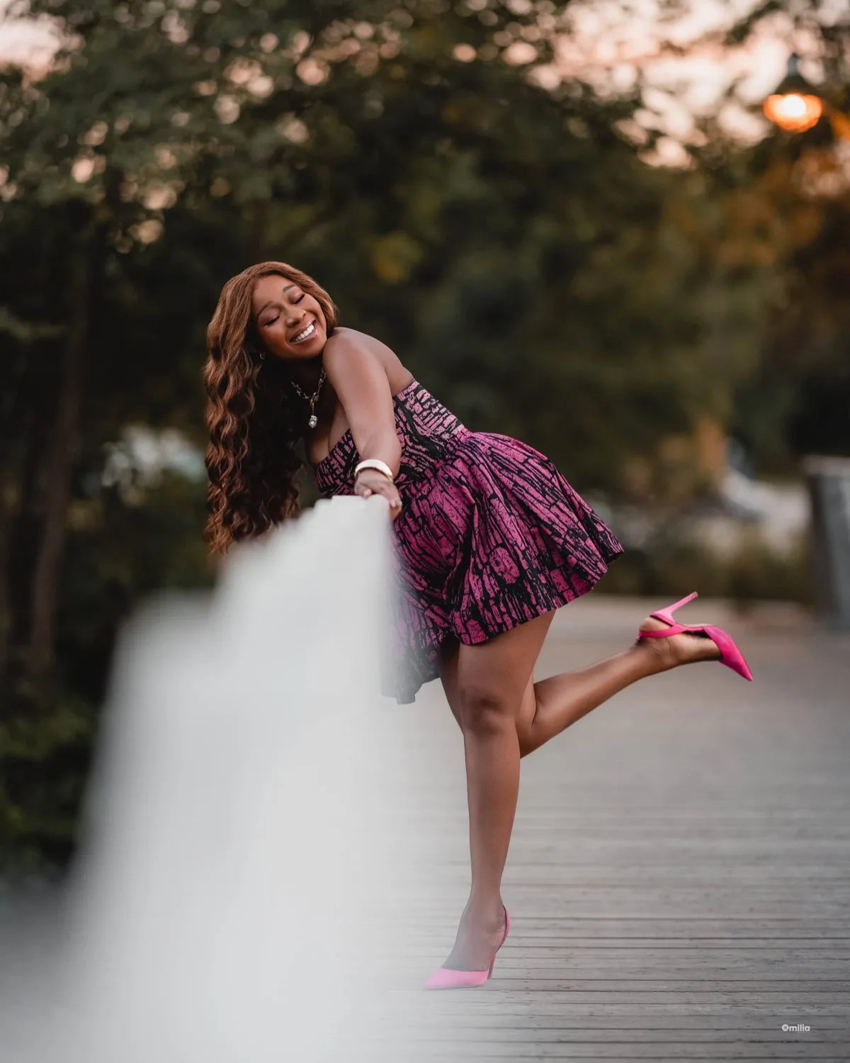 Laughing woman in pink patterned dress and heels leaning on white railing at dusk