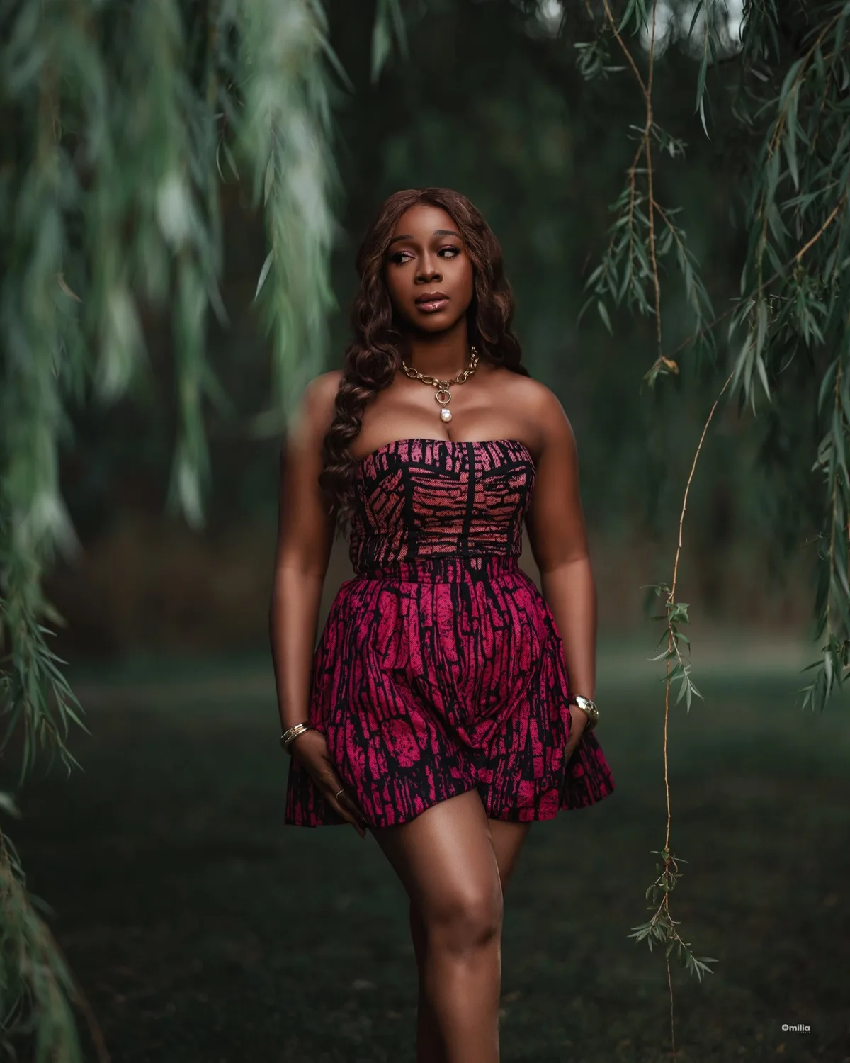 Woman in pink and black patterned strapless dress standing among draping willow branches outdoors