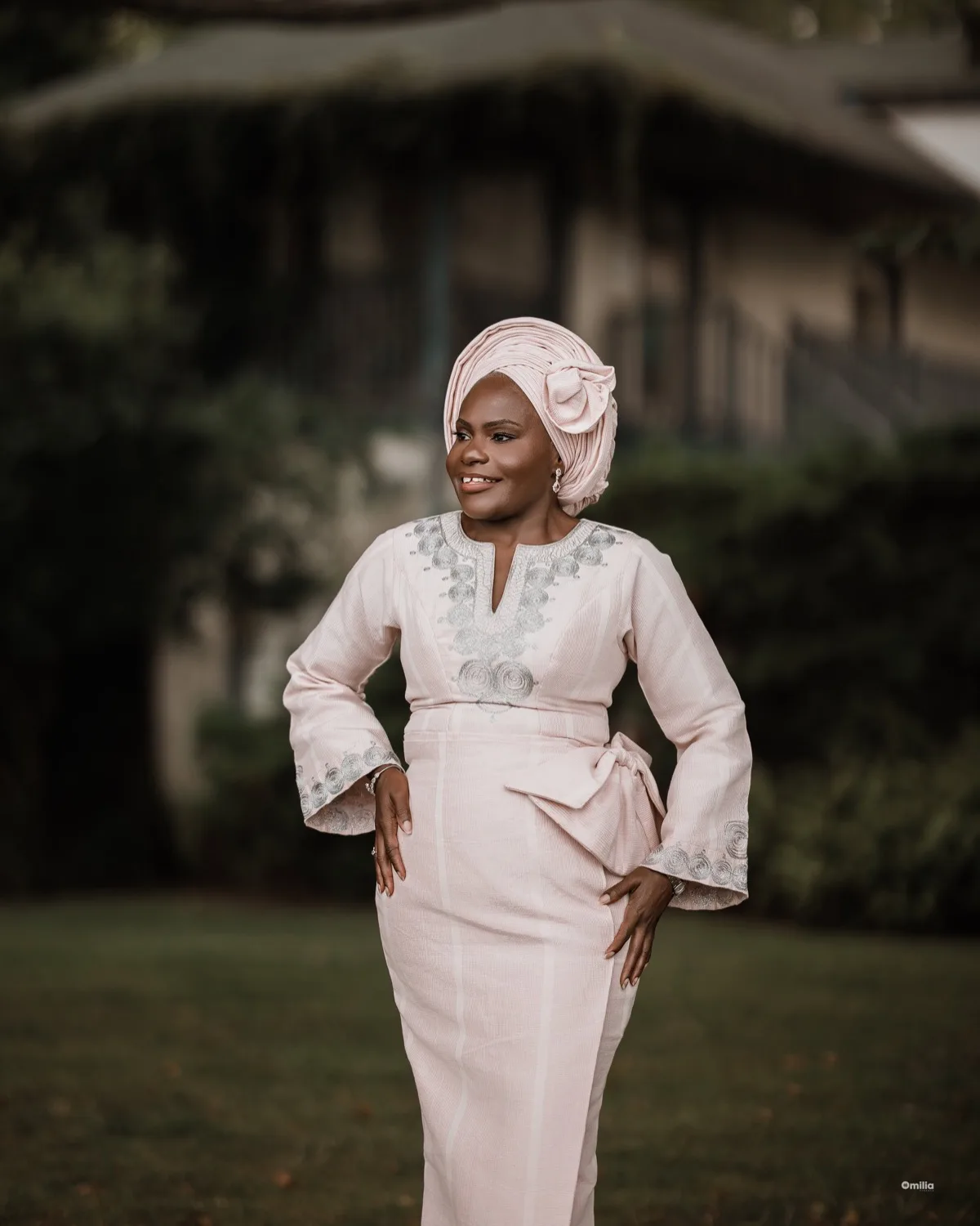Smiling woman in pale pink embroidered outfit and head wrap posing outdoors against blurred greenery