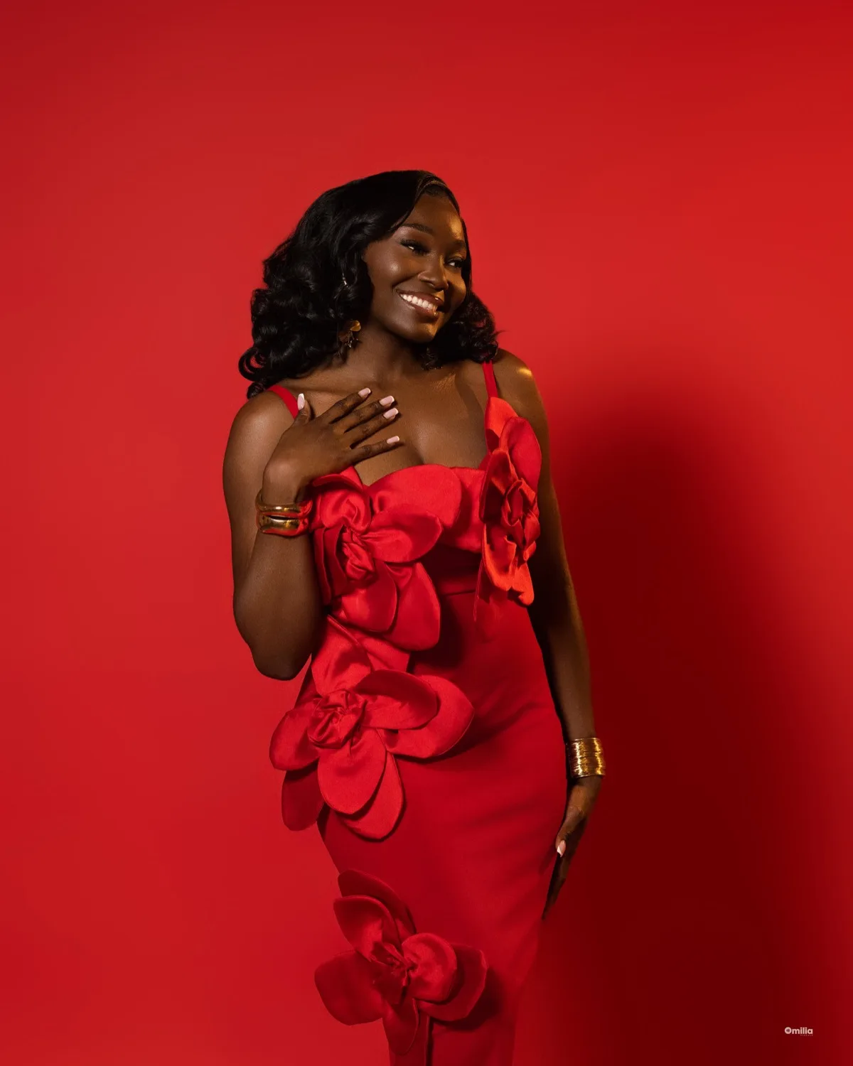 Smiling woman in red gown with floral rose appliqués and gold bangles against bold red backdrop