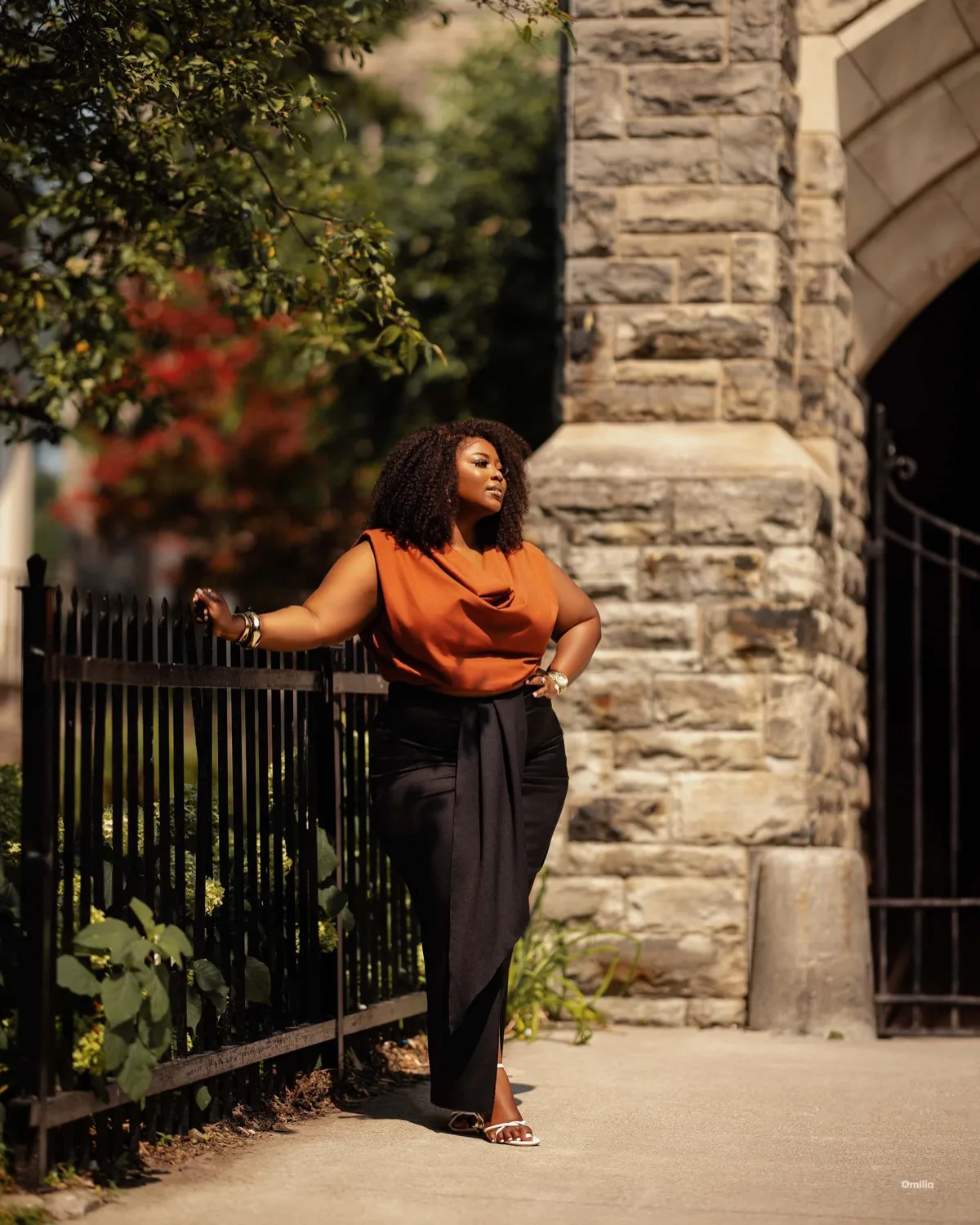 Woman with afro in rust blouse and black trousers standing by iron fence near stone archway