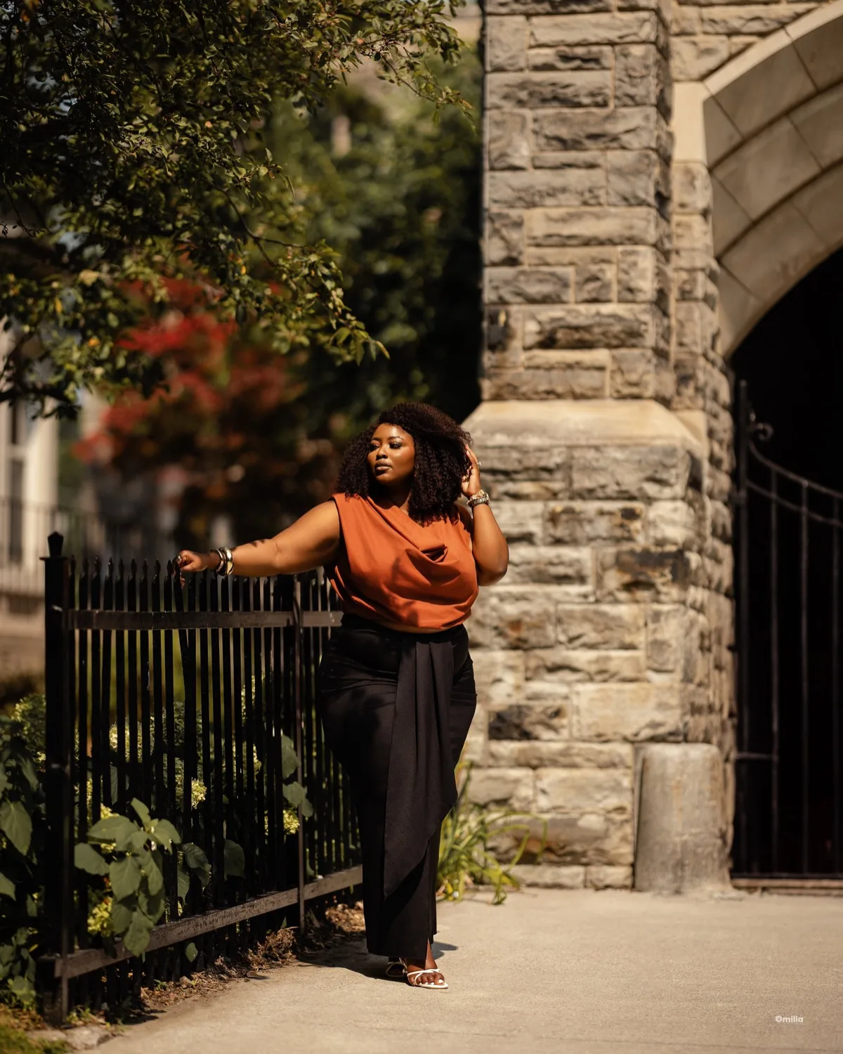 Woman in rust top and black trousers leaning on an iron fence by a stone building