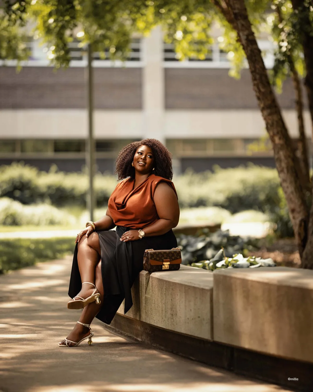 Woman with afro in rust sleeveless top seated on stone ledge in sunlit courtyard with trees