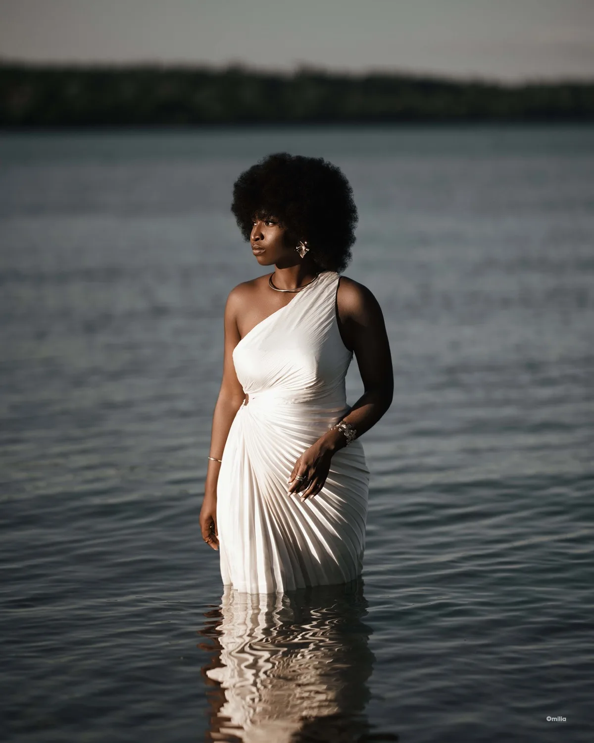 Woman with afro in one-shoulder white pleated dress standing in calm lake water at dusk