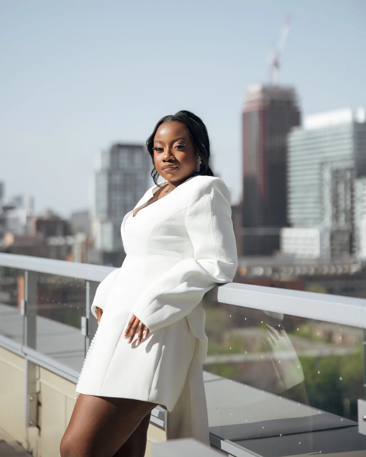 Woman in white blazer dress leaning on a rooftop glass railing with a city skyline behind