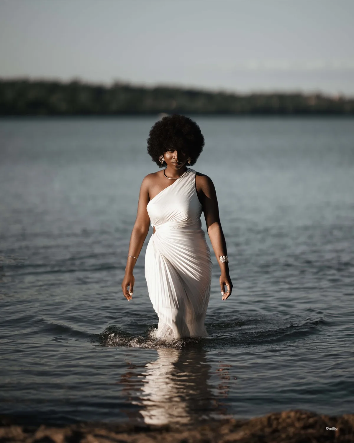 Woman with afro in white one-shoulder dress wading through lake water with tree-lined horizon behind