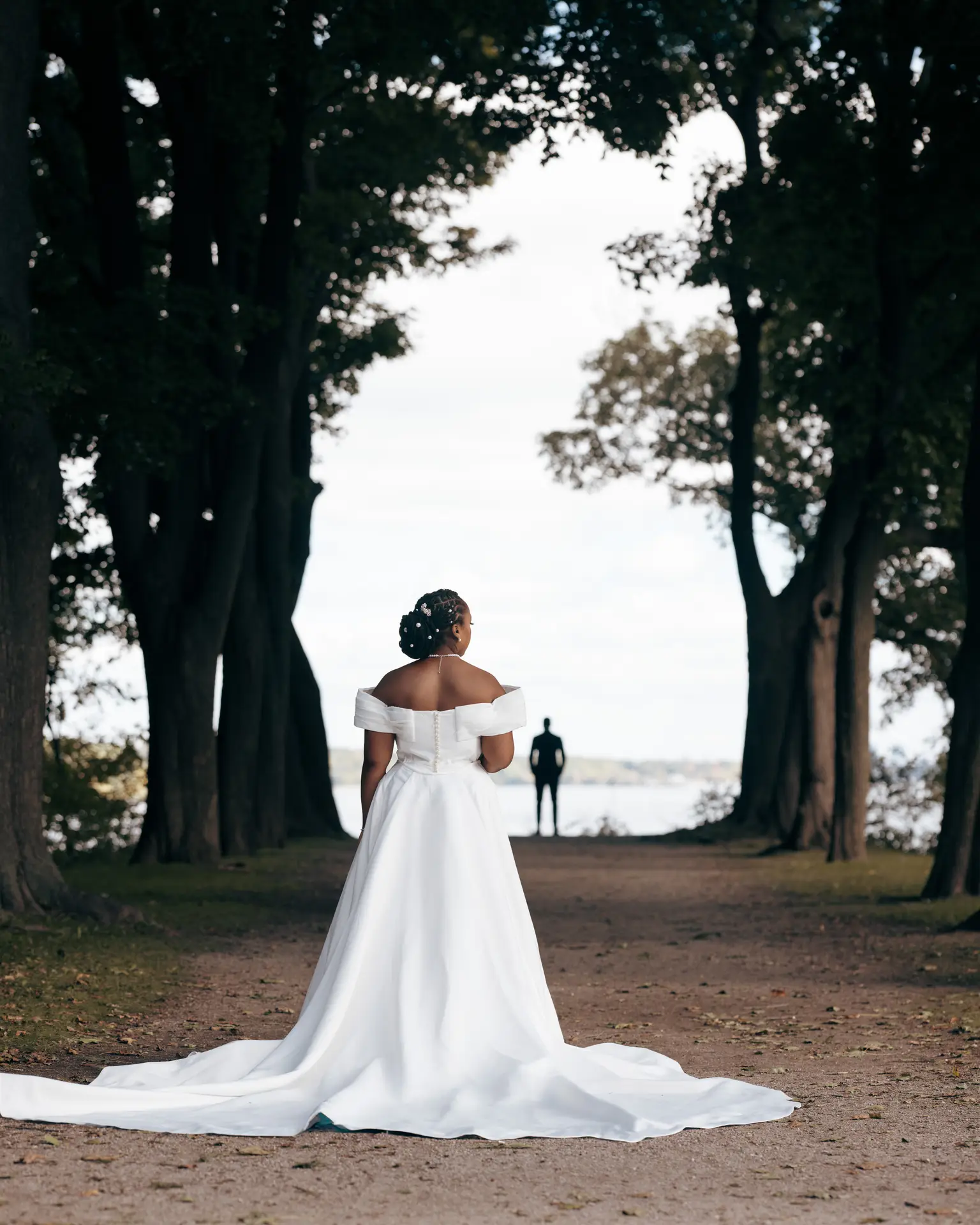 Bride in off-shoulder gown facing a distant groom silhouetted between tall trees by water