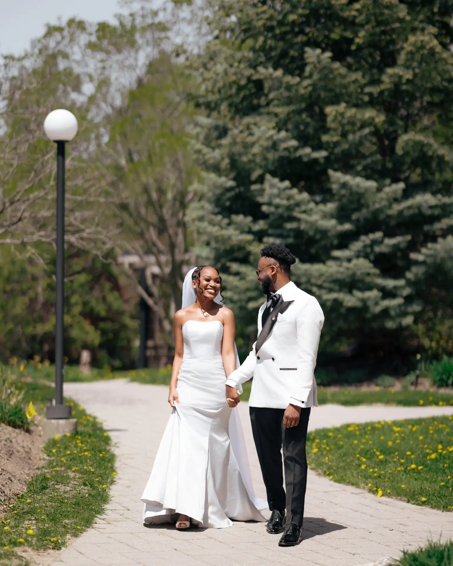 Bride and groom holding hands strolling a sunlit garden path past a lamppost