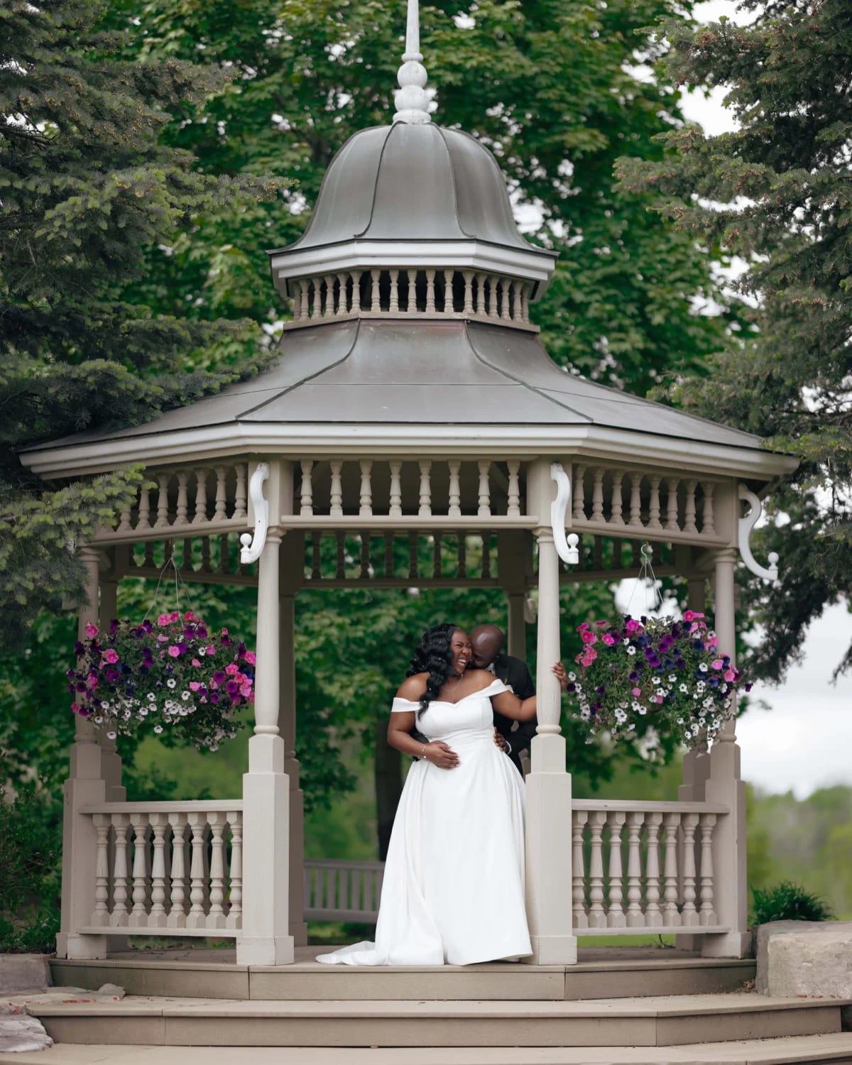 Bride and groom embracing beneath a gazebo with hanging flower baskets and trees behind