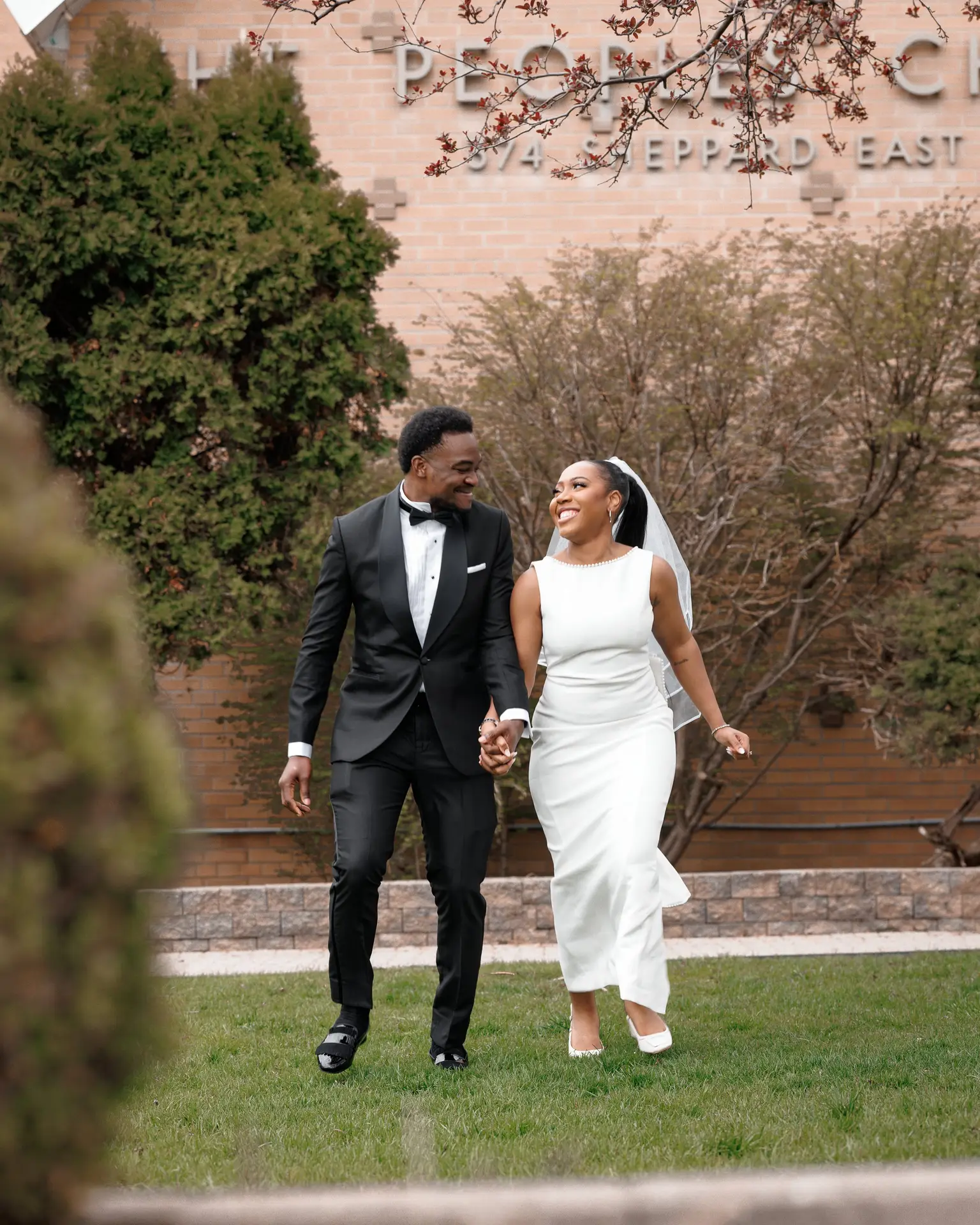 Bride and groom holding hands strolling across a lawn outside a brick building