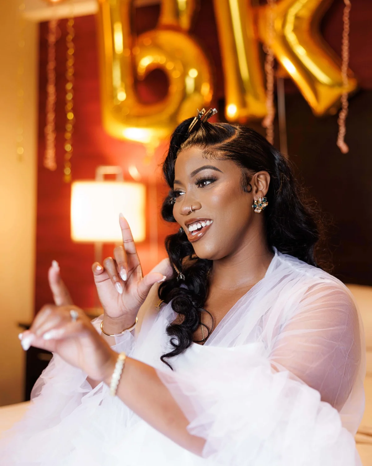 Smiling bride in sheer tulle robe gesturing playfully beneath gold letter balloons
