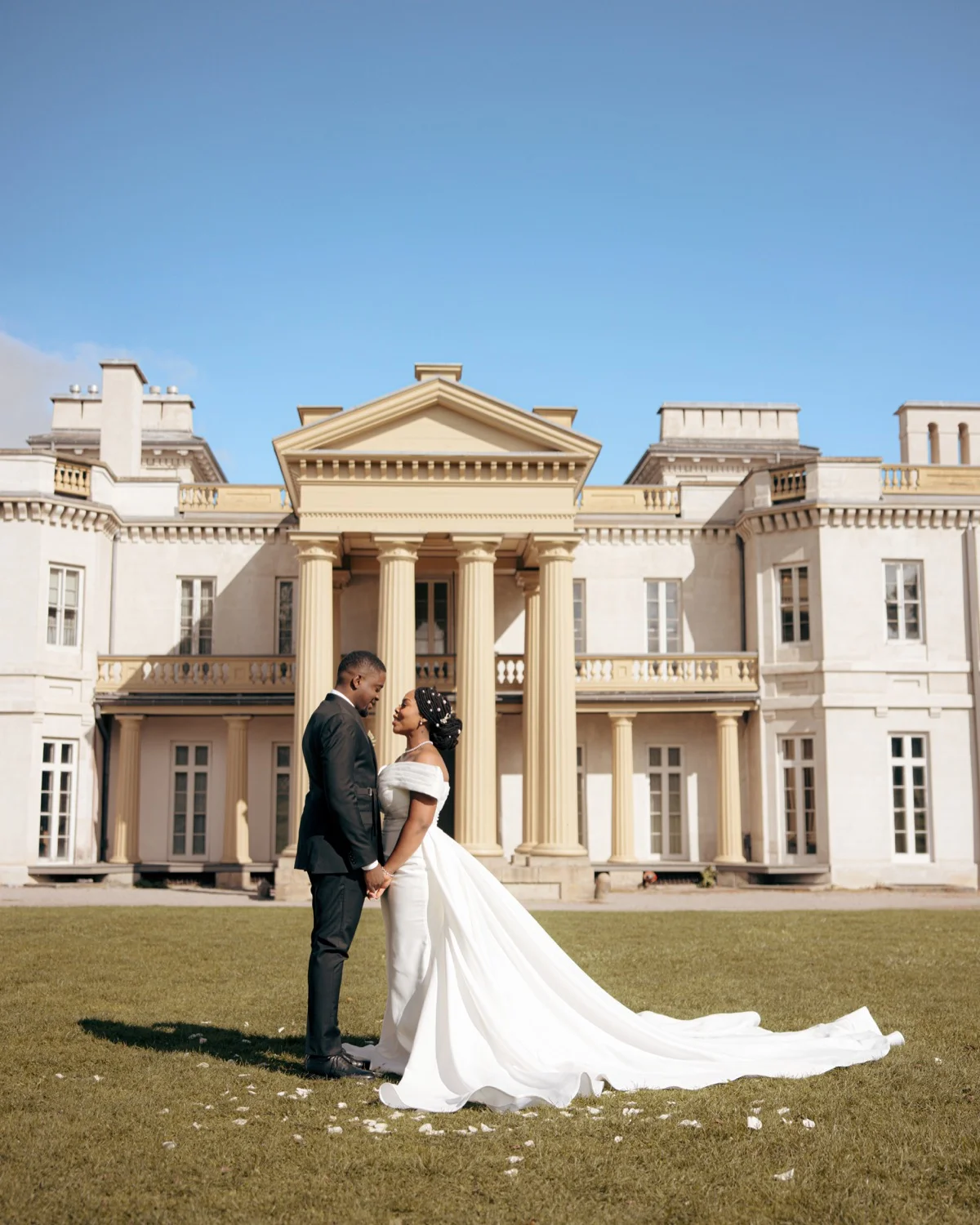 Bride and groom holding hands on a lawn before a columned mansion, train fanned out
