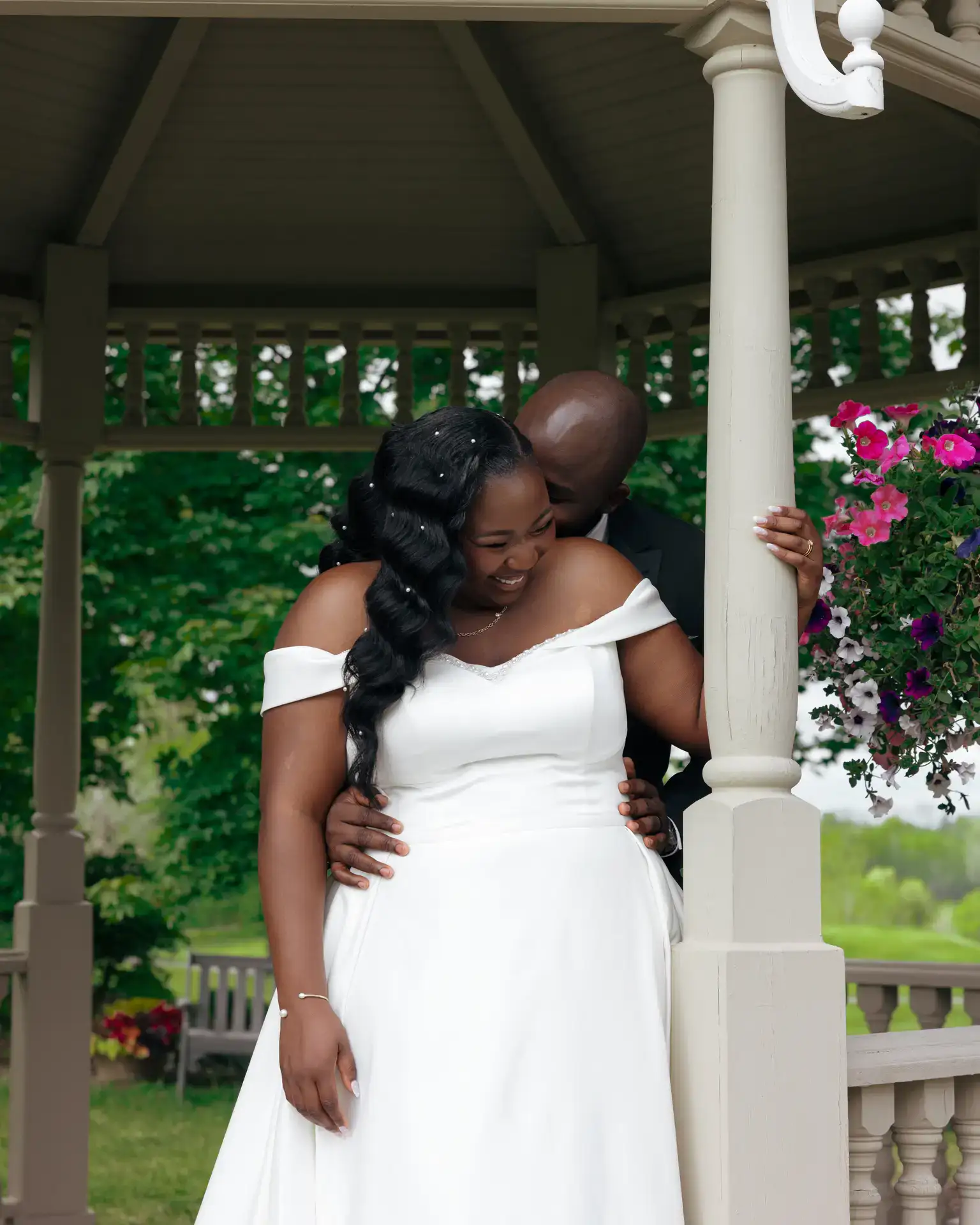 Groom kissing laughing bride's cheek from behind beneath a gazebo with hanging petunias