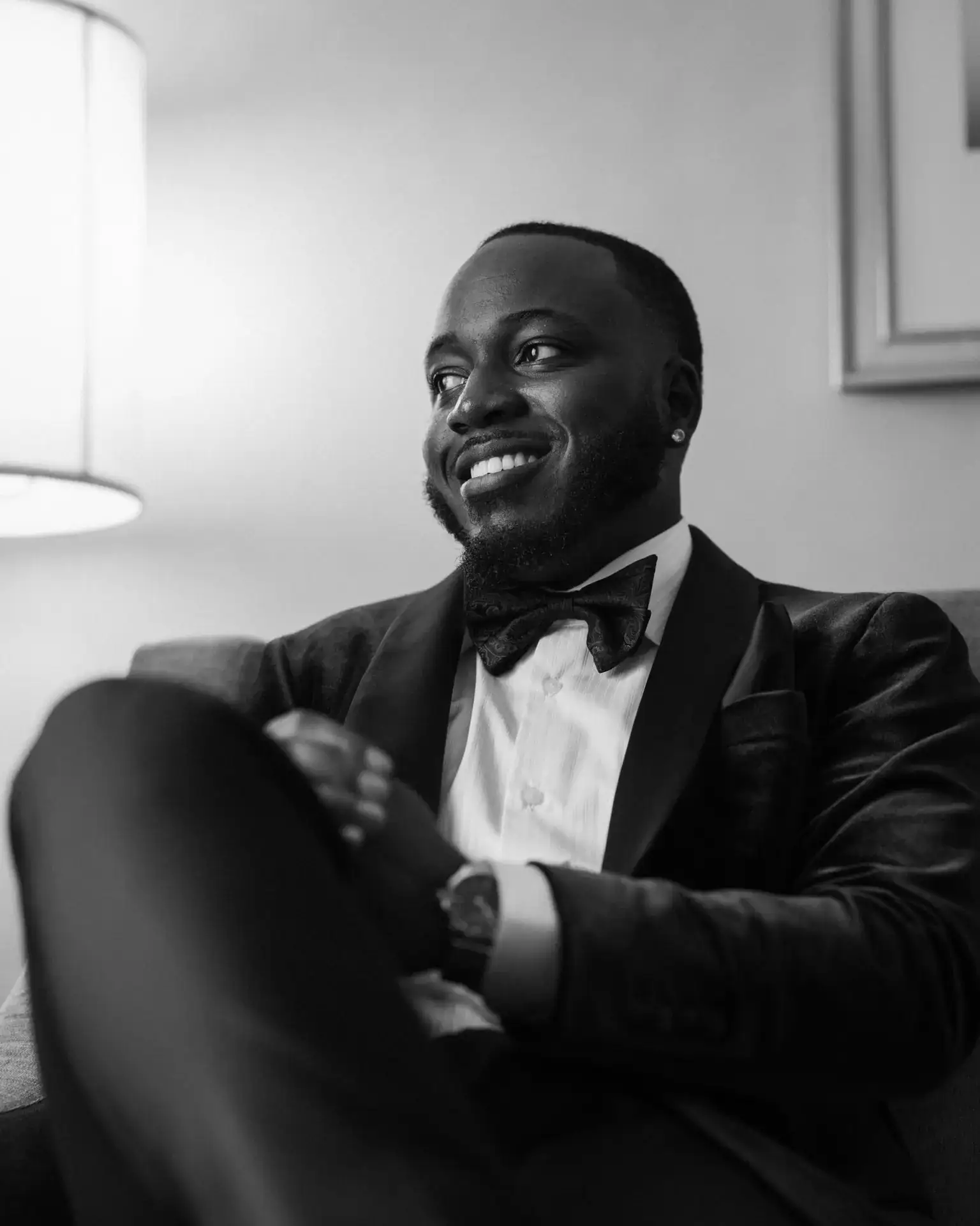 Groom in velvet tuxedo and paisley bow tie smiling seated in an armchair