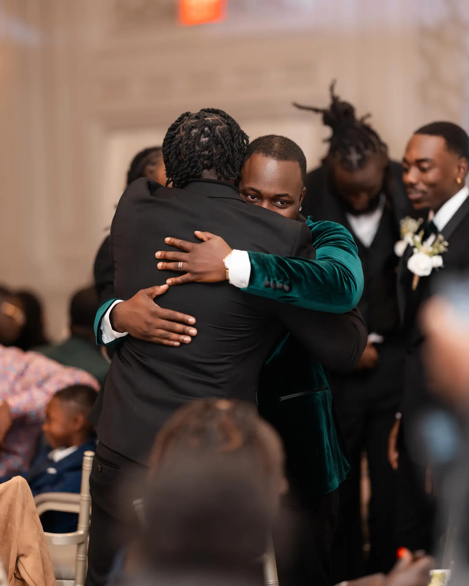 Groom in green velvet jacket embracing a guest amid a crowded reception celebration