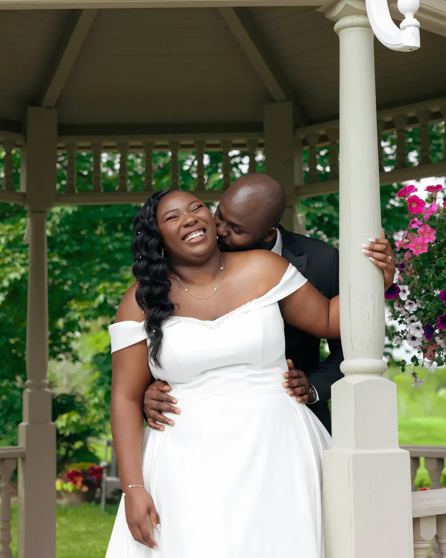 Groom kissing the laughing bride's cheek from behind beside a gazebo post with petunias