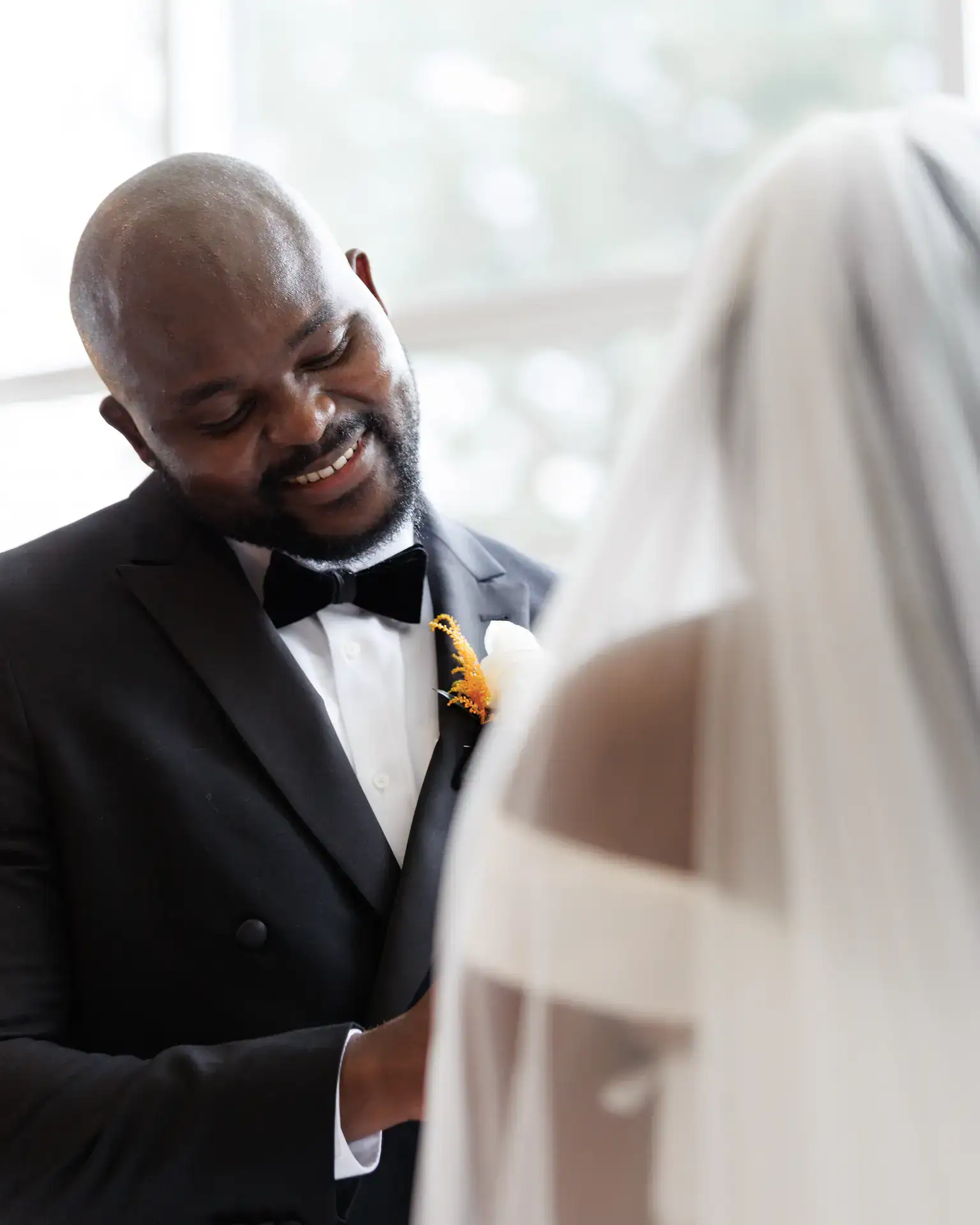 Groom in black tuxedo smiling warmly at his veiled bride beside a bright window