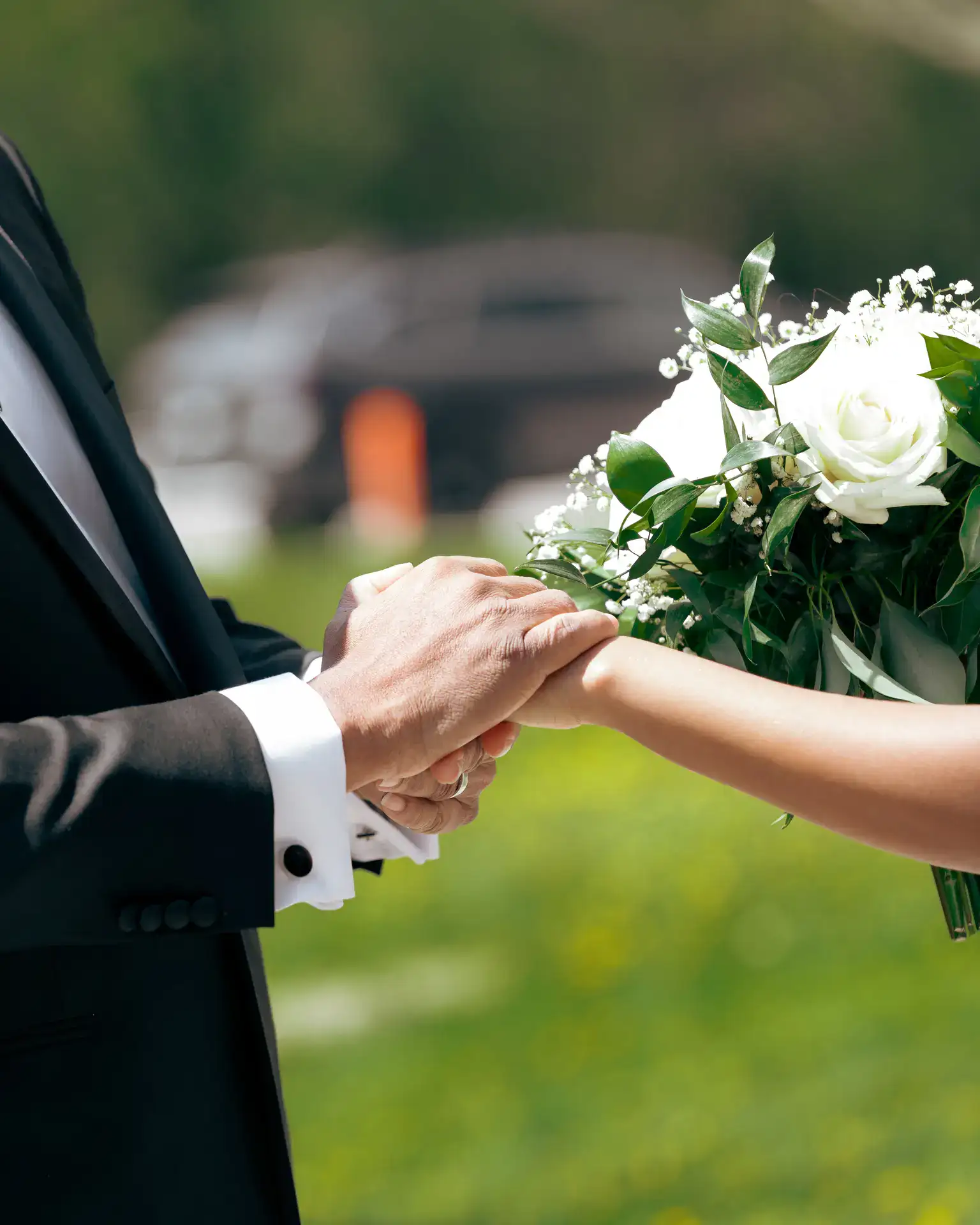 Suited man clasping bride's hand beside white rose bouquet on sunlit lawn