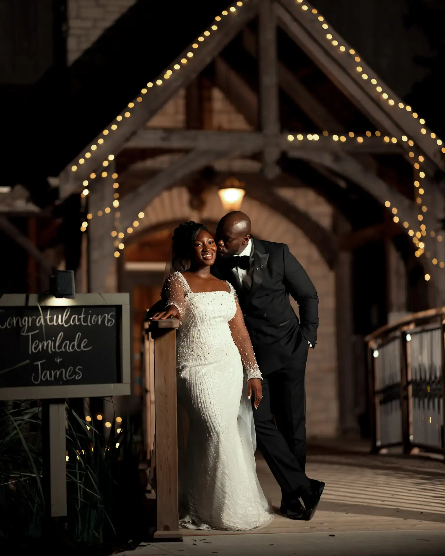 Groom kissing the bride's cheek at night beside a chalkboard sign under string lights