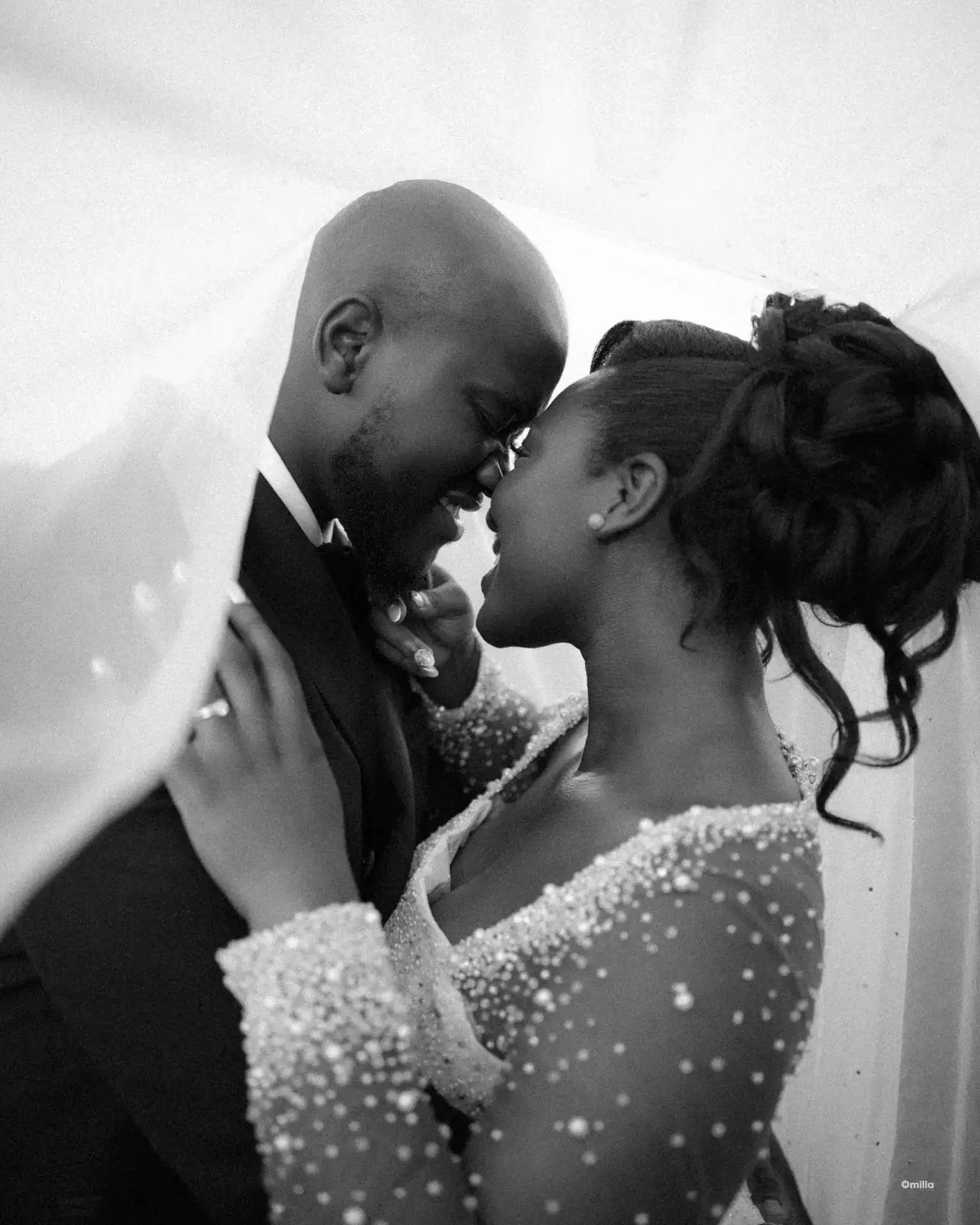 Couple touching noses beneath the bride's veil, her beaded sleeves catching the light