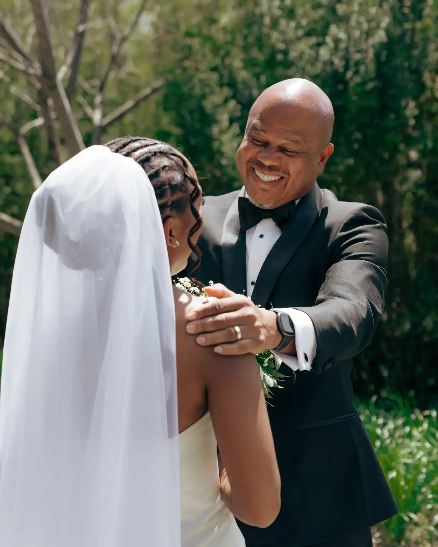 Smiling older man in tuxedo resting hand on veiled bride's shoulder amid greenery
