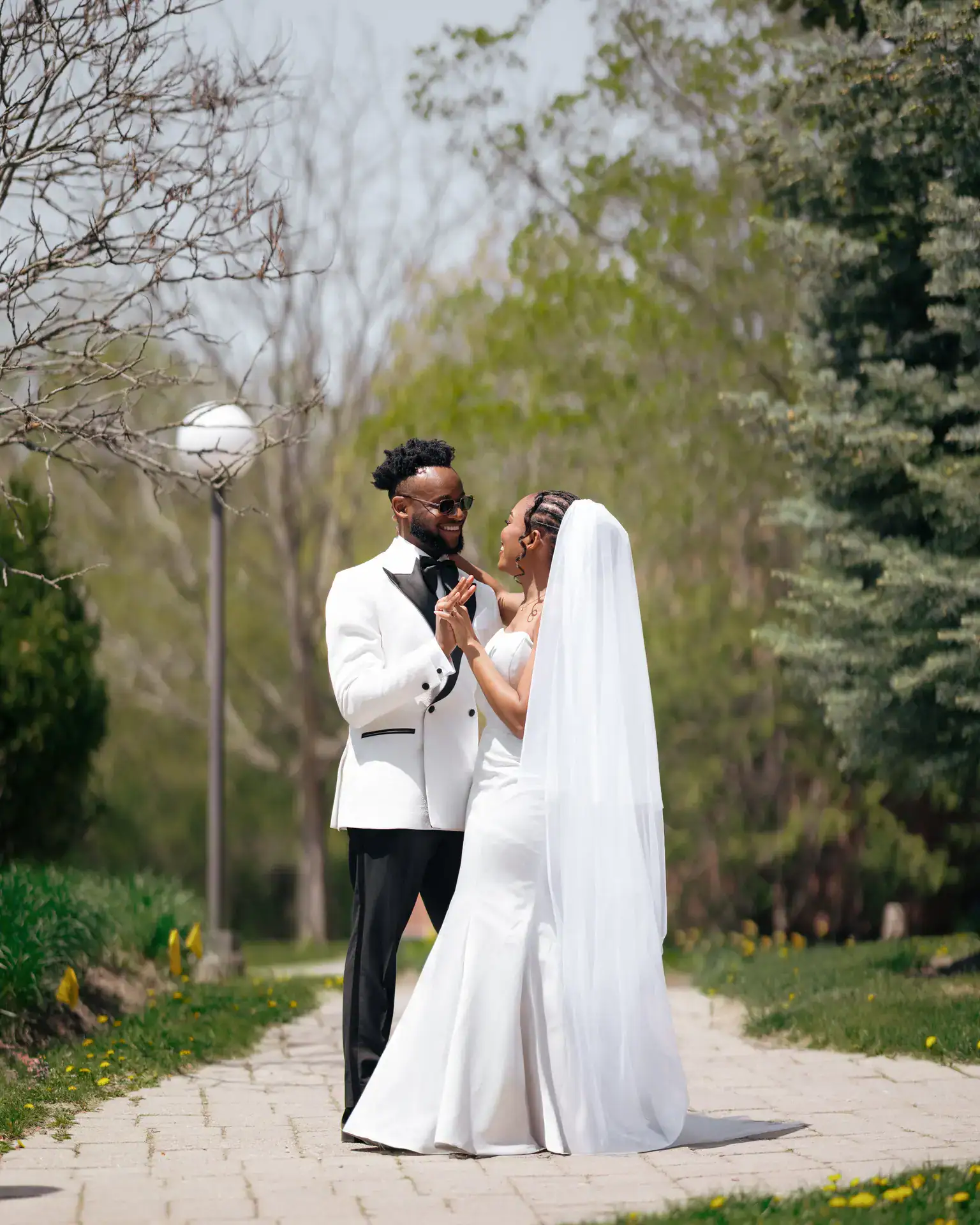 Groom in white dinner jacket and veiled bride clasping hands on a garden path