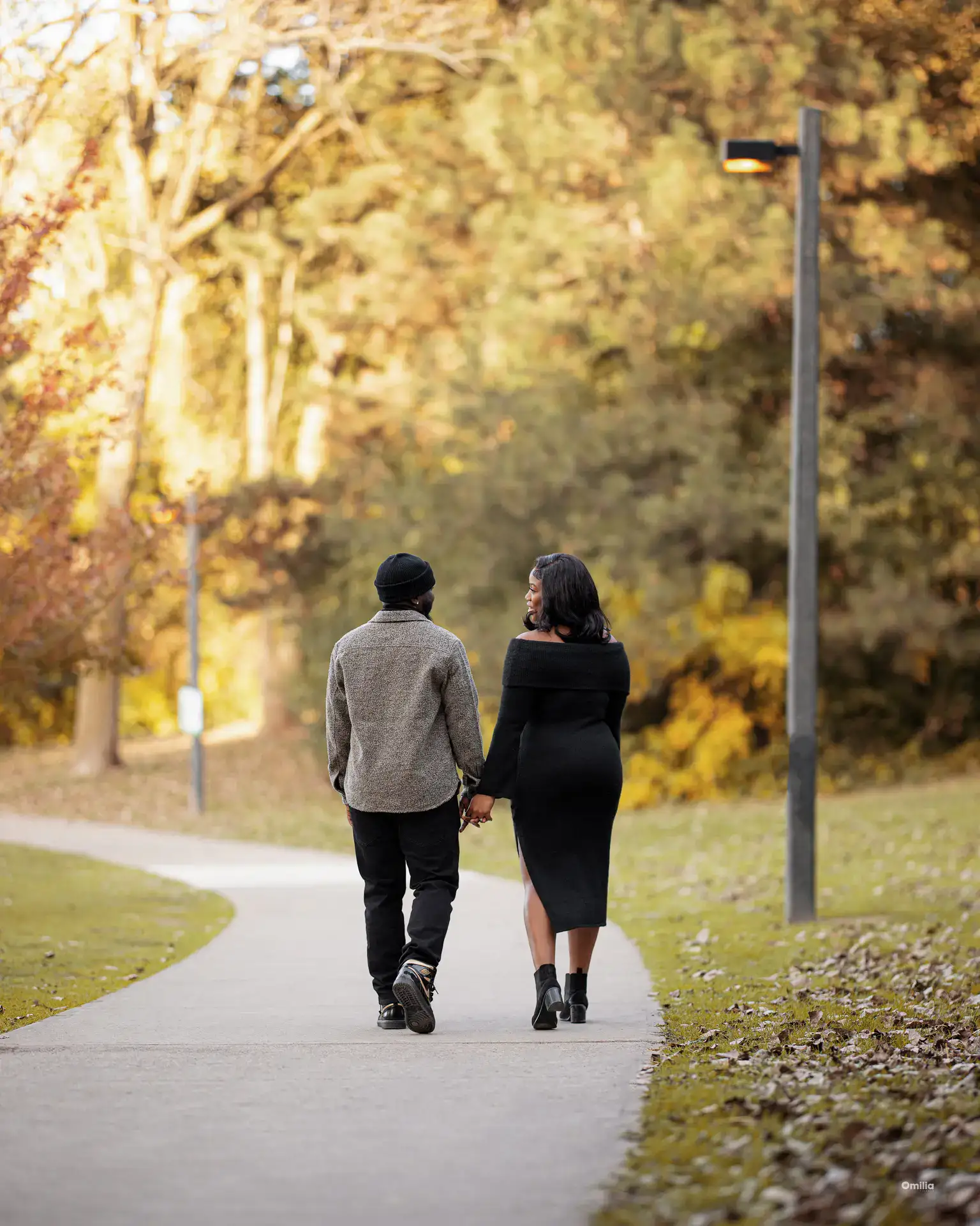 Couple walking hand in hand along a paved park path beneath autumn trees