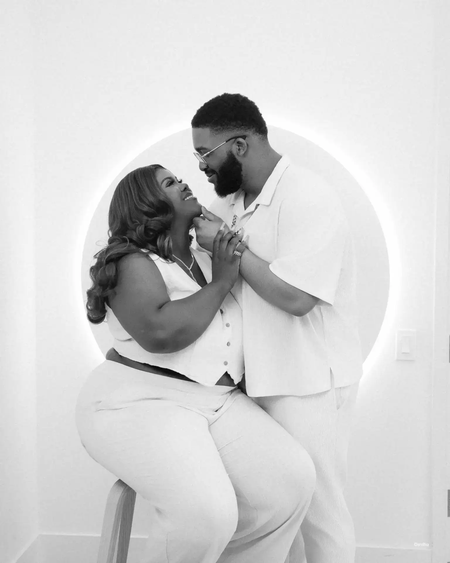Couple in matching white outfits posing before a backlit circular alcove, man lifting her chin