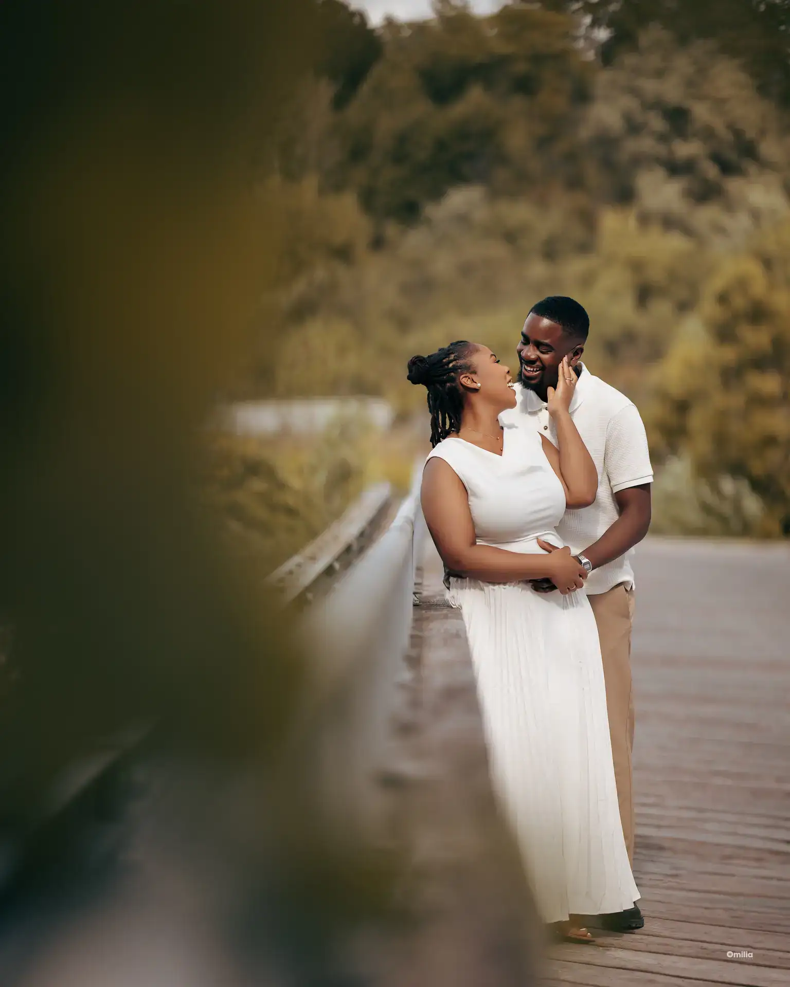 Couple embracing on a boardwalk bridge, woman in white dress touching partner's cheek