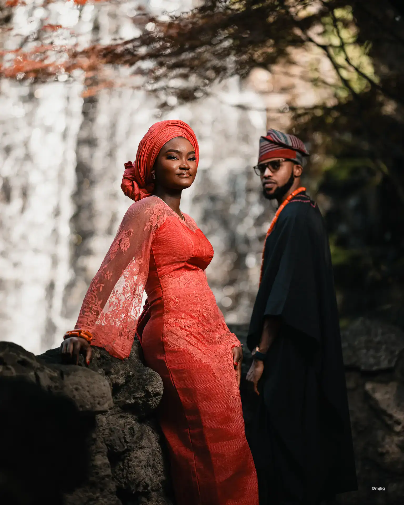 Woman in coral lace dress and gele posing by a waterfall, partner in dark agbada behind