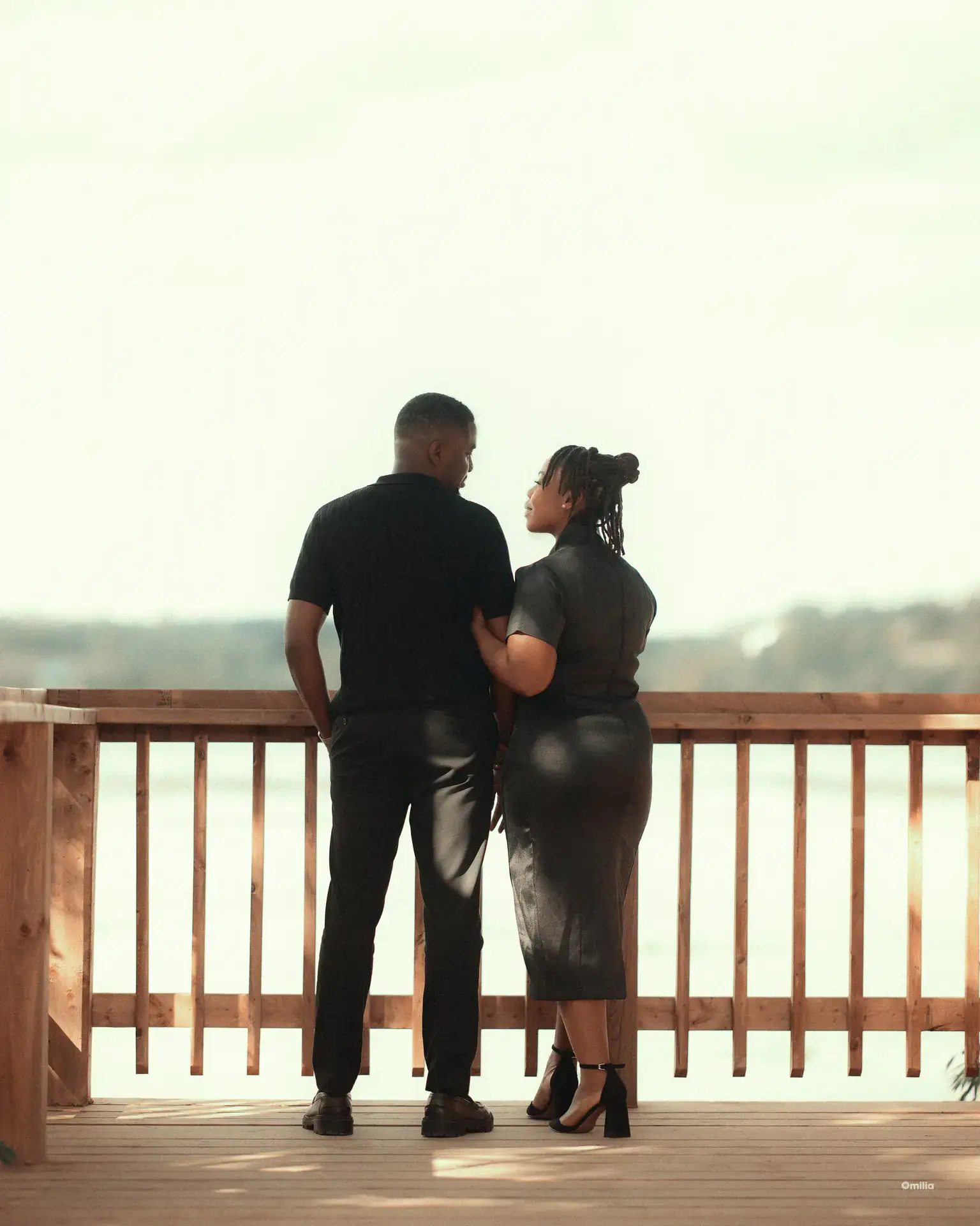 Couple in black outfits standing at a wooden deck railing overlooking water