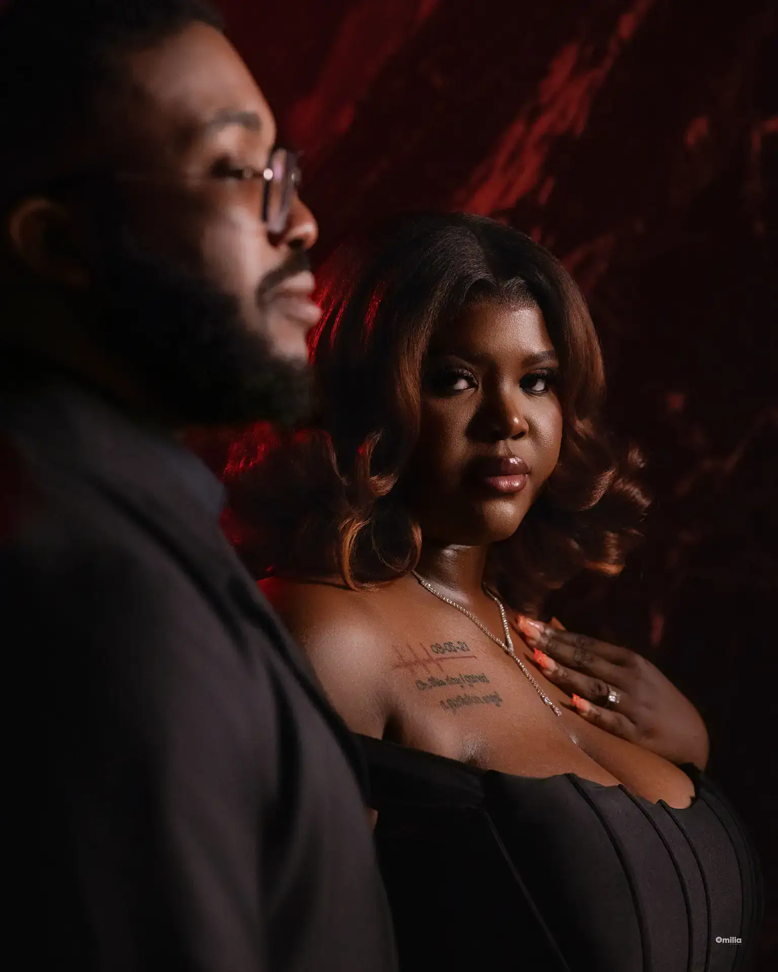 Woman resting hand on chest beside partner's blurred profile against red-lit backdrop