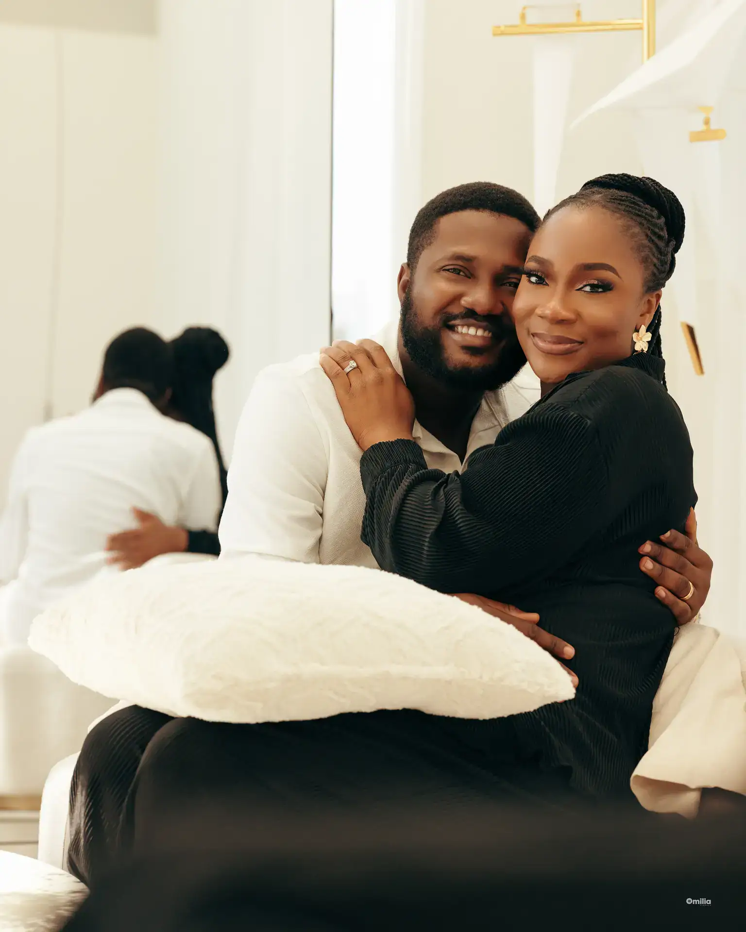 Couple seated in an embrace holding a plush white pillow, mirror reflection visible behind