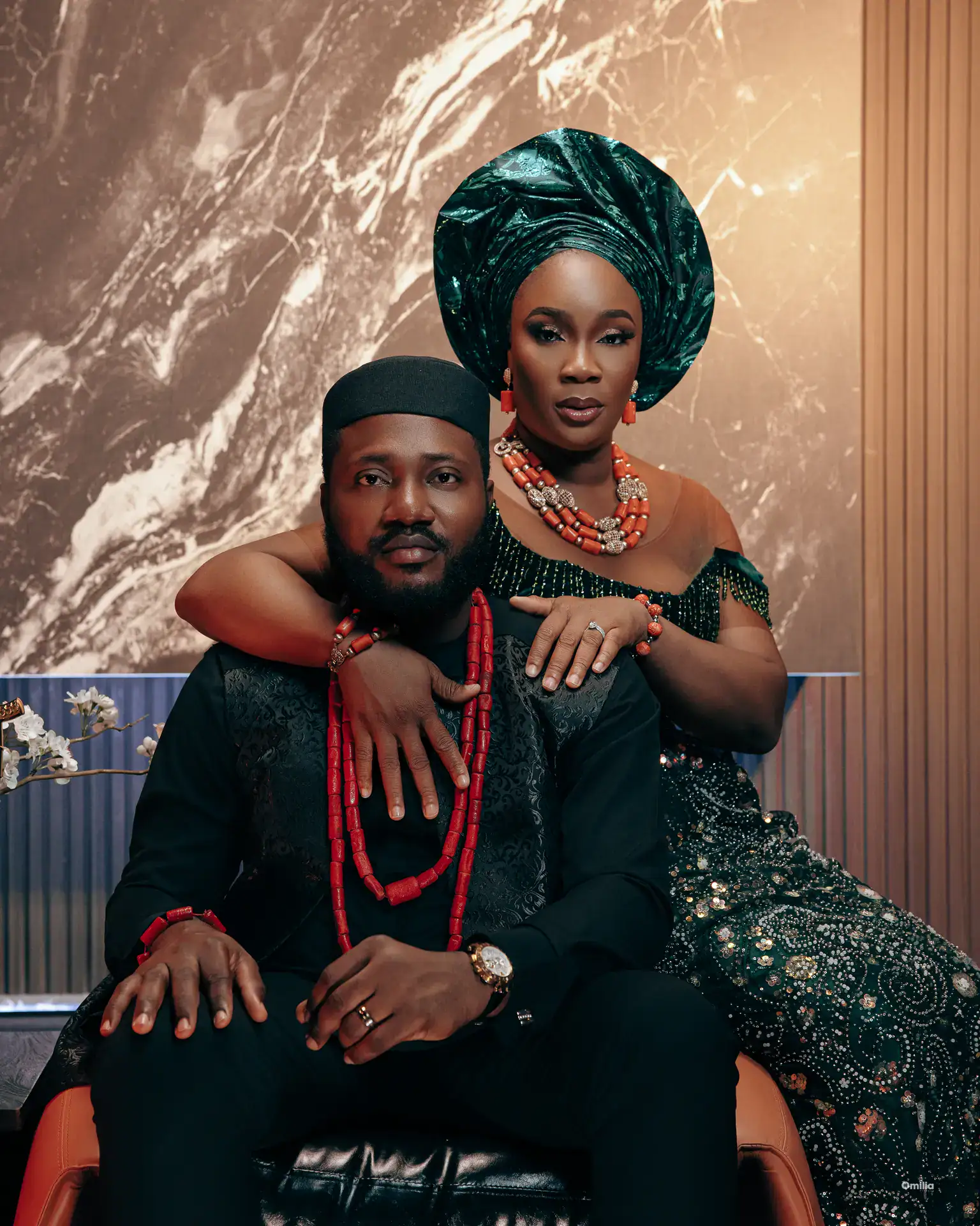 Couple in traditional attire with coral beads, woman in emerald gele seated against marble backdrop
